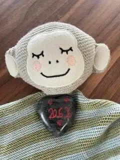 Soft plush monkey toy with closed eyes and pink cheeks, wearing a dark heart-shaped pendant with red numbers around its neck on a striped blanket.
