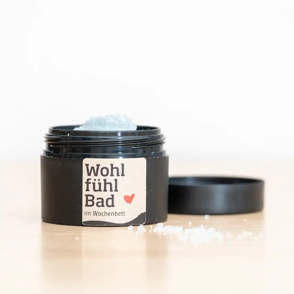 Open black jar of bath salts with blue crystals; label reads 'Wohl fühl Bad im Wochenbett' on a wooden surface with a loose crystal nearby.