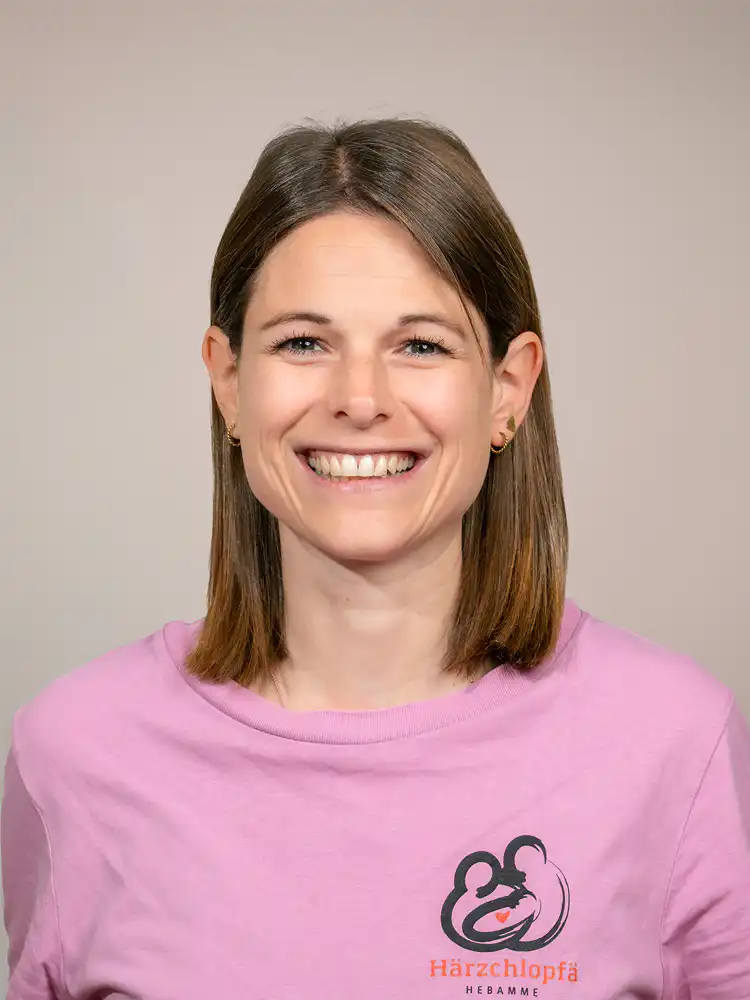 Portrait of a smiling woman with shoulder-length brown hair wearing a pink shirt with a left-chest logo against a neutral background.