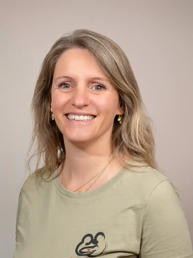 Portrait of a smiling woman with shoulder-length blond hair, wearing a beige T-shirt with a small logo against a neutral background.