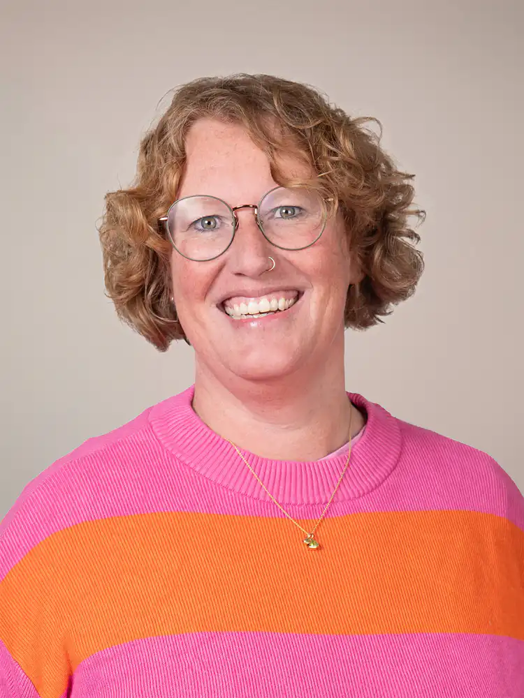 Smiling woman with short curly hair, round glasses, and a pink-and-orange striped sweater wearing a small gold necklace.