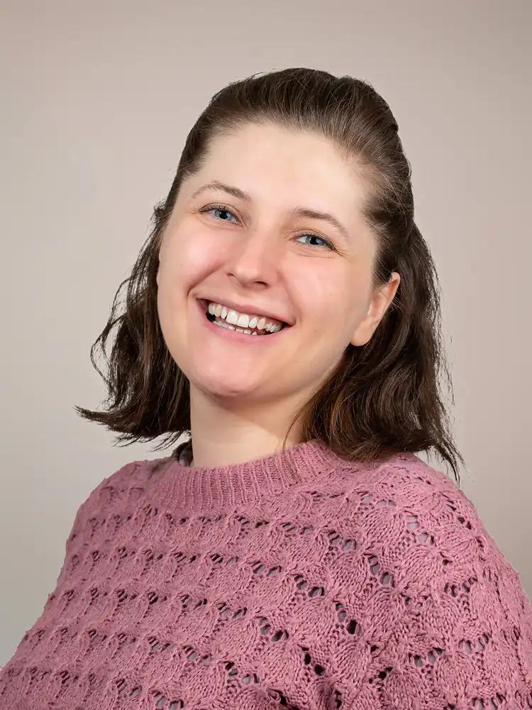 Smiling woman with shoulder-length dark hair wearing a pink knitted sweater against a neutral background.