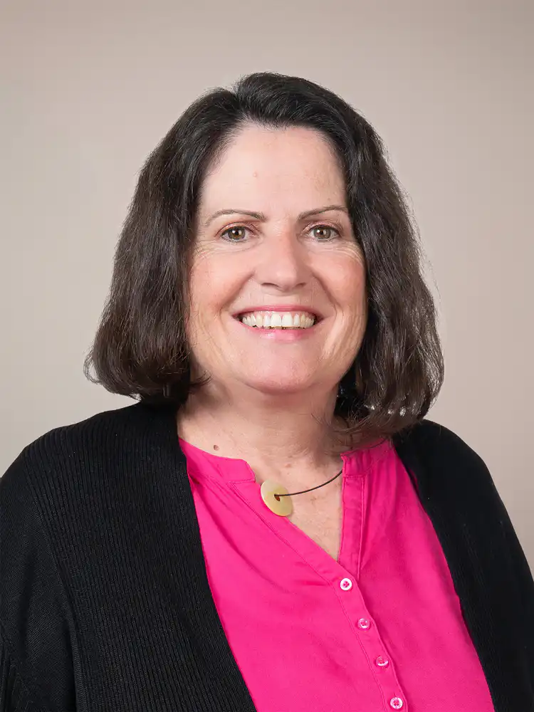 Portrait of a smiling woman with dark hair, wearing a pink blouse and black cardigan against a neutral beige background, looking at the camera.