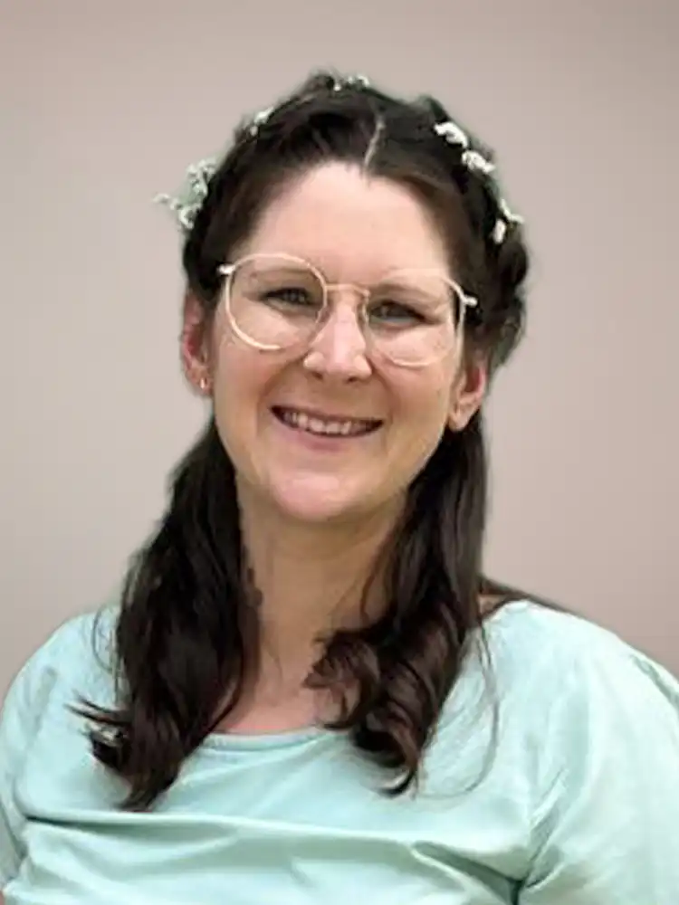 Smiling woman with round glasses, dark hair adorned with small white flowers, wearing a light blue top.