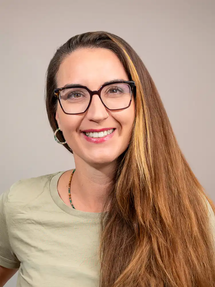 Smiling woman with long light brown hair, black glasses, hoop earrings, wearing a pale green shirt.