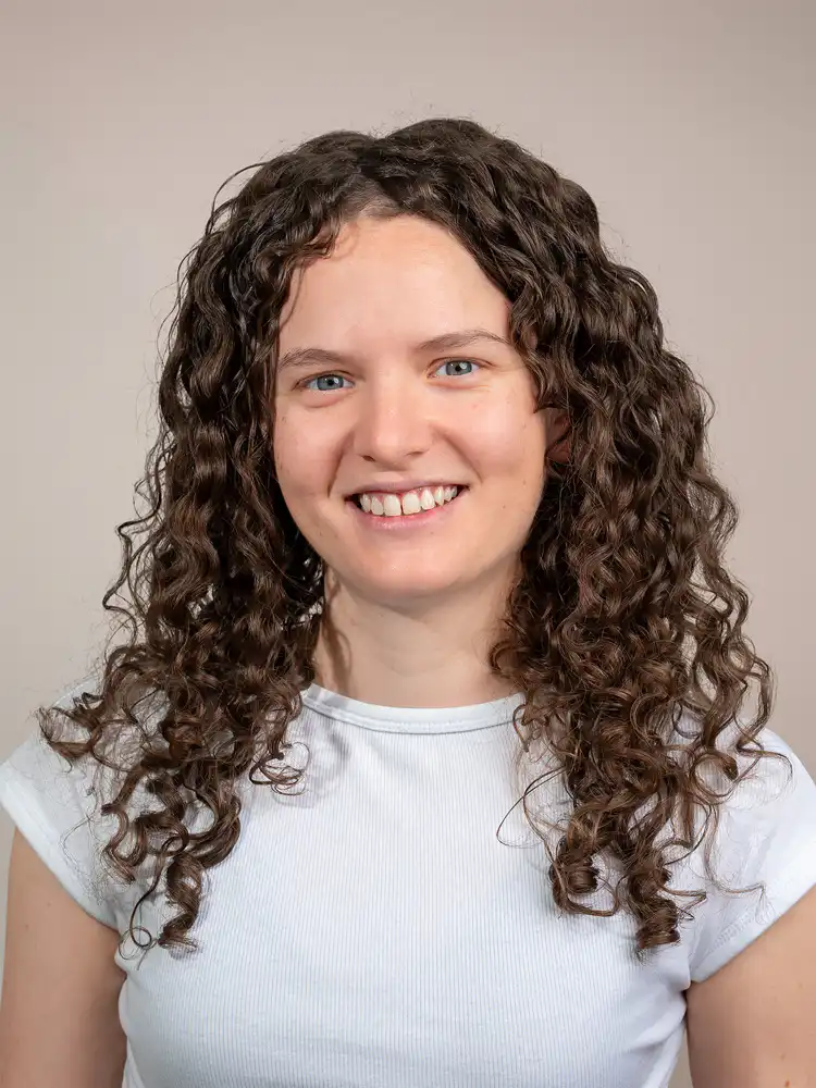 Smiling young woman with long curly brown hair wearing a white ribbed shirt against a neutral background