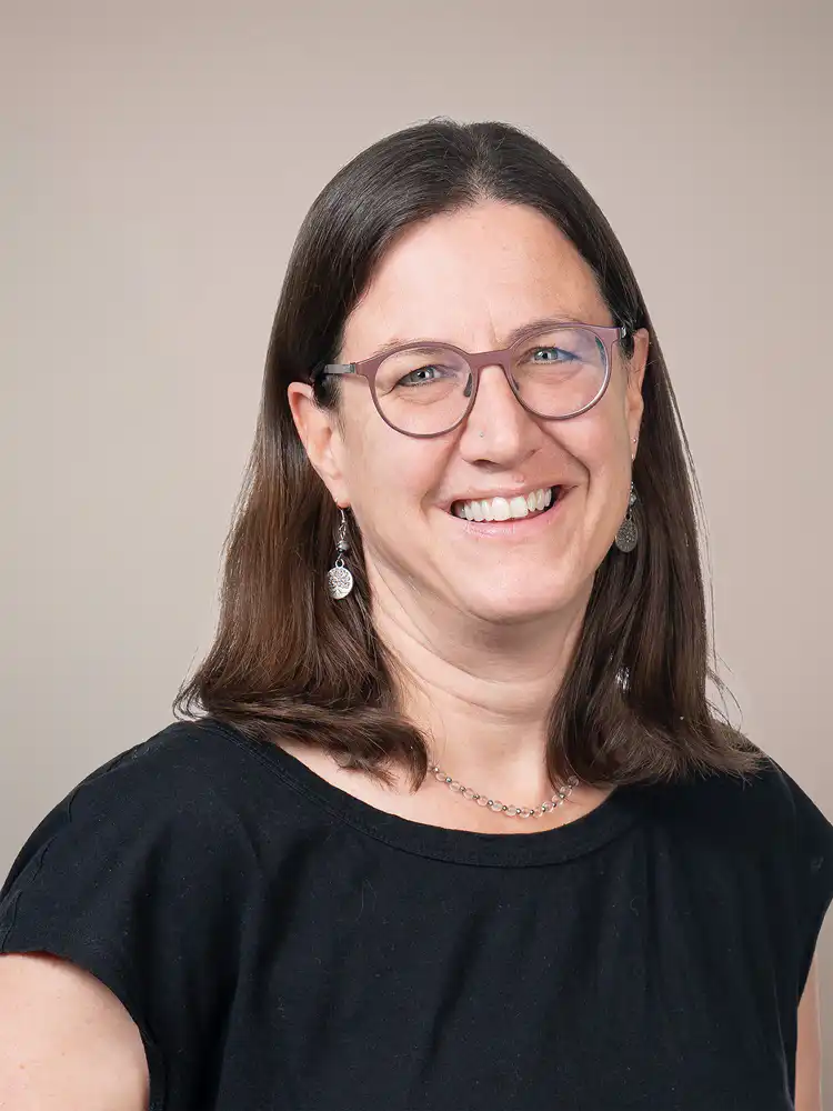 Portrait of a smiling woman with shoulder-length brown hair and round glasses, wearing a black top and beaded necklace against a beige background in a professional headshot.