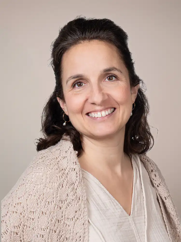 Smiling woman with dark brown hair wearing a beige knitted cardigan and pearl earrings, posed against a neutral beige backdrop.