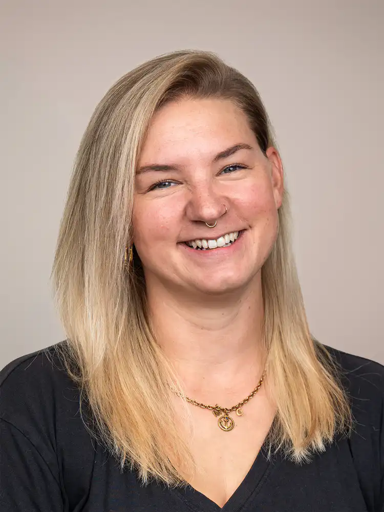 Smiling woman with shoulder-length blonde hair, wearing a nose ring and gold necklace against a neutral background.