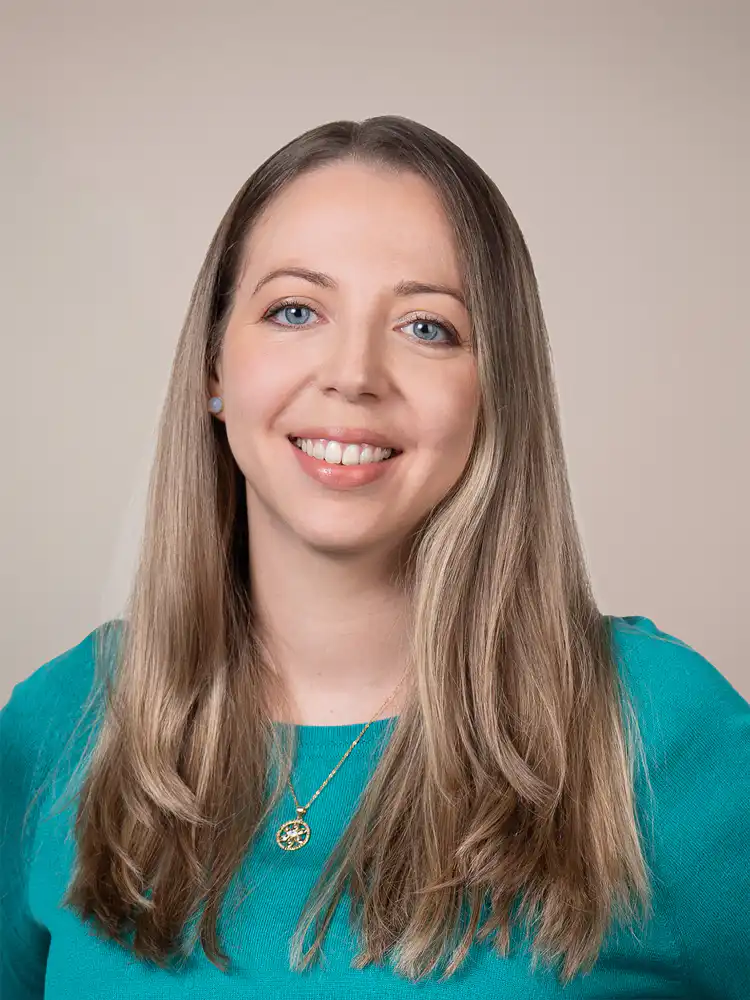 Smiling woman with long light brown hair in a teal top, wearing a gold pendant necklace, against a neutral backdrop.