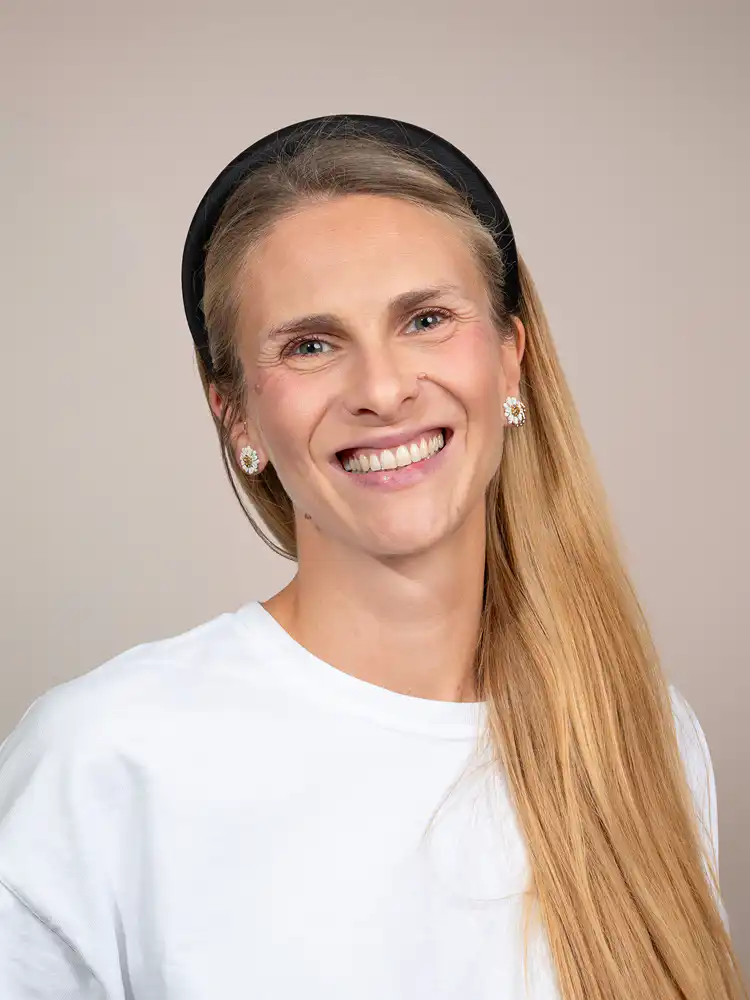 Smiling woman with long blonde hair wearing a black headband and white shirt, floral earrings visible on each ear