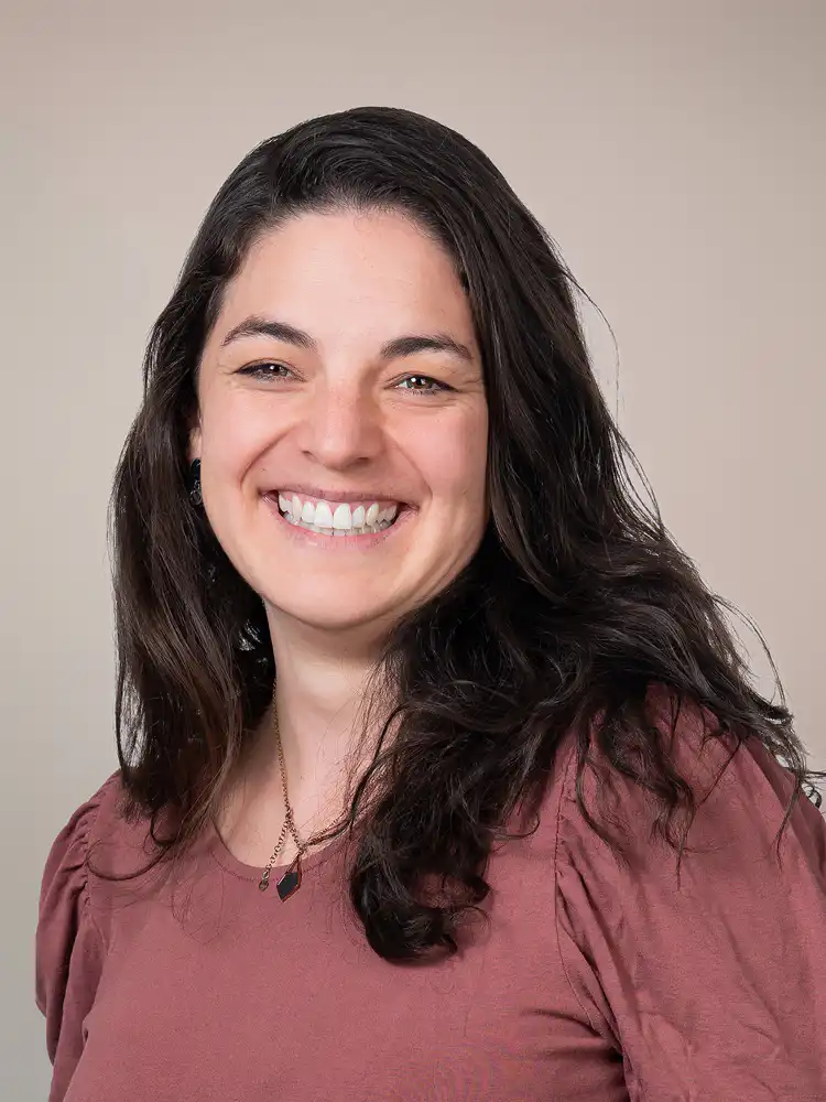 Portrait of a smiling woman with long dark hair in a pink blouse against a neutral backdrop.