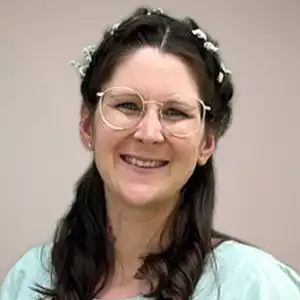 Smiling woman with round glasses and a floral headband, wearing a light blue top against a neutral background.