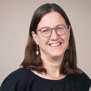 Smiling woman with shoulder-length brown hair, glasses, and dangling earrings, wearing a black top.