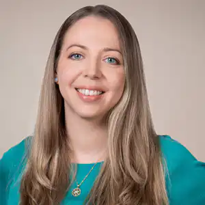 Portrait of a smiling woman with long light-brown hair, wearing a teal blouse and a gold pendant necklace.